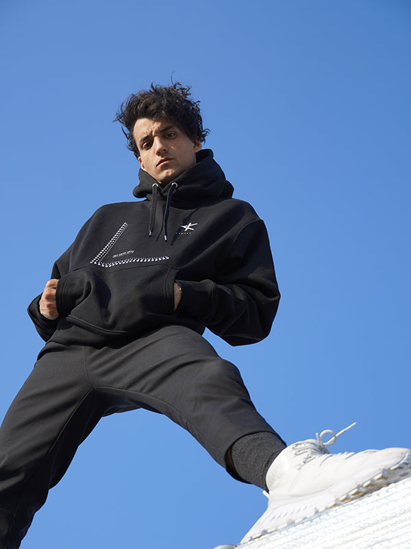 Person wearing a black hoodie and pants with white shoes against a clear blue sky.